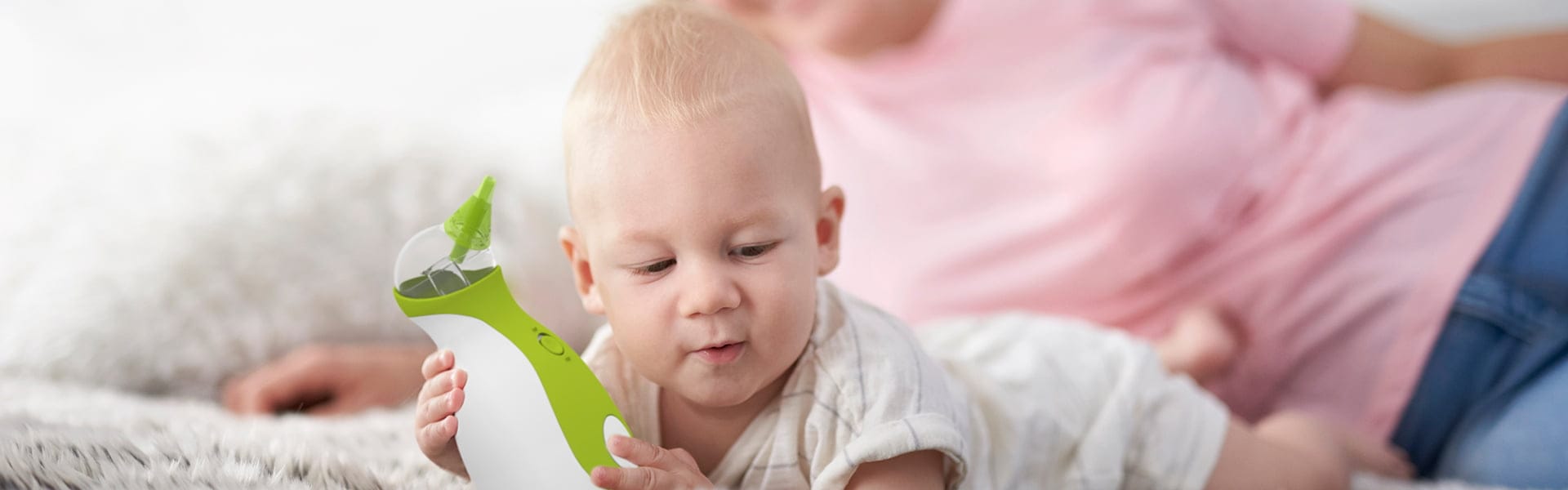 Ein Baby liegt auf seinem Bauch auf einem Teppich und hält den Nosiboo Go tragbaren Baby Nasensauger in seinen Händen. Im Hintergrund sichtbar: die Silhouetten seiner Mutter