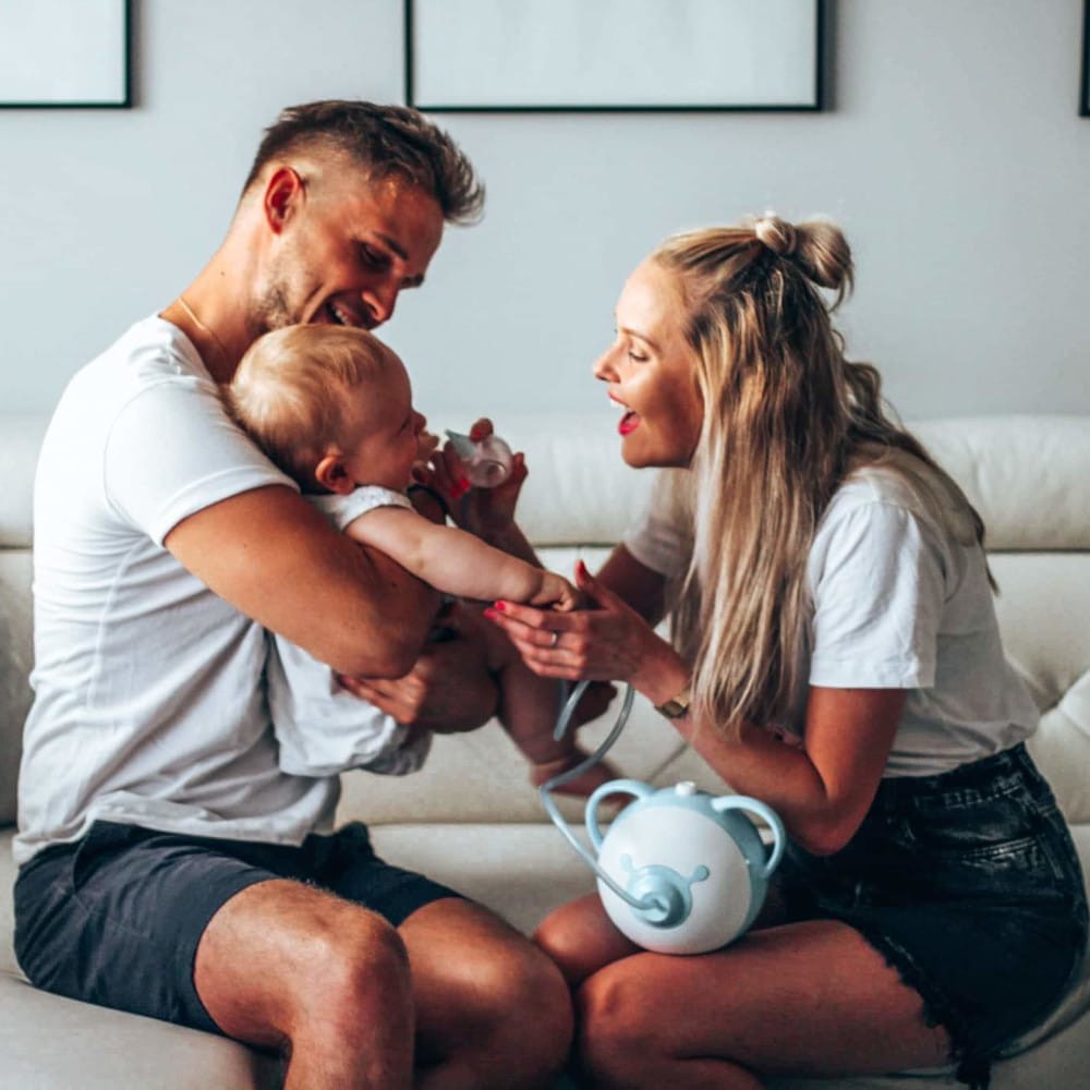 Mutter und Vater saugen gemeinsam die Nase ihres Babys mit einem elektrischen Nosiboo Pro Nasensauger ab. Die Farben des Bildes wirken magazinhaft oberflächlich wie etwa aus den 80er Jahren. Die Eltern tragen shorts und T-Shirts. Alle haben große Freude an der Sache.