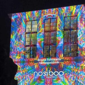 Das Nosiboo-Logo mit bunten Lichten an der Wand eines Gebäudes, die über die Förderung "Bronze-Stufe" des Pécser Zsolnay-Lichtfestivals informiert.