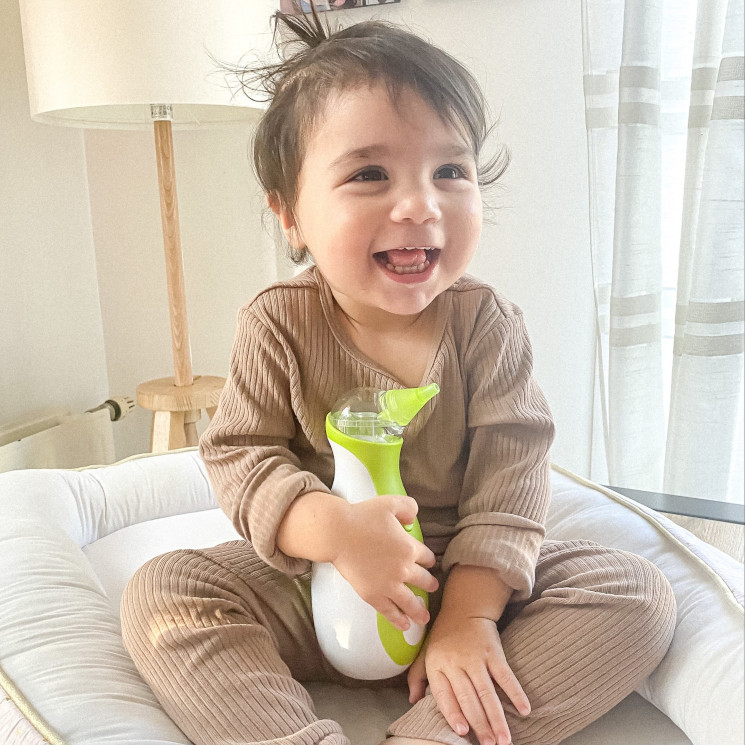 A laughing little boy sitting in his pyjamas in the children's room and holding the Nosiboo Go portable nasal aspirator to clean little noses.