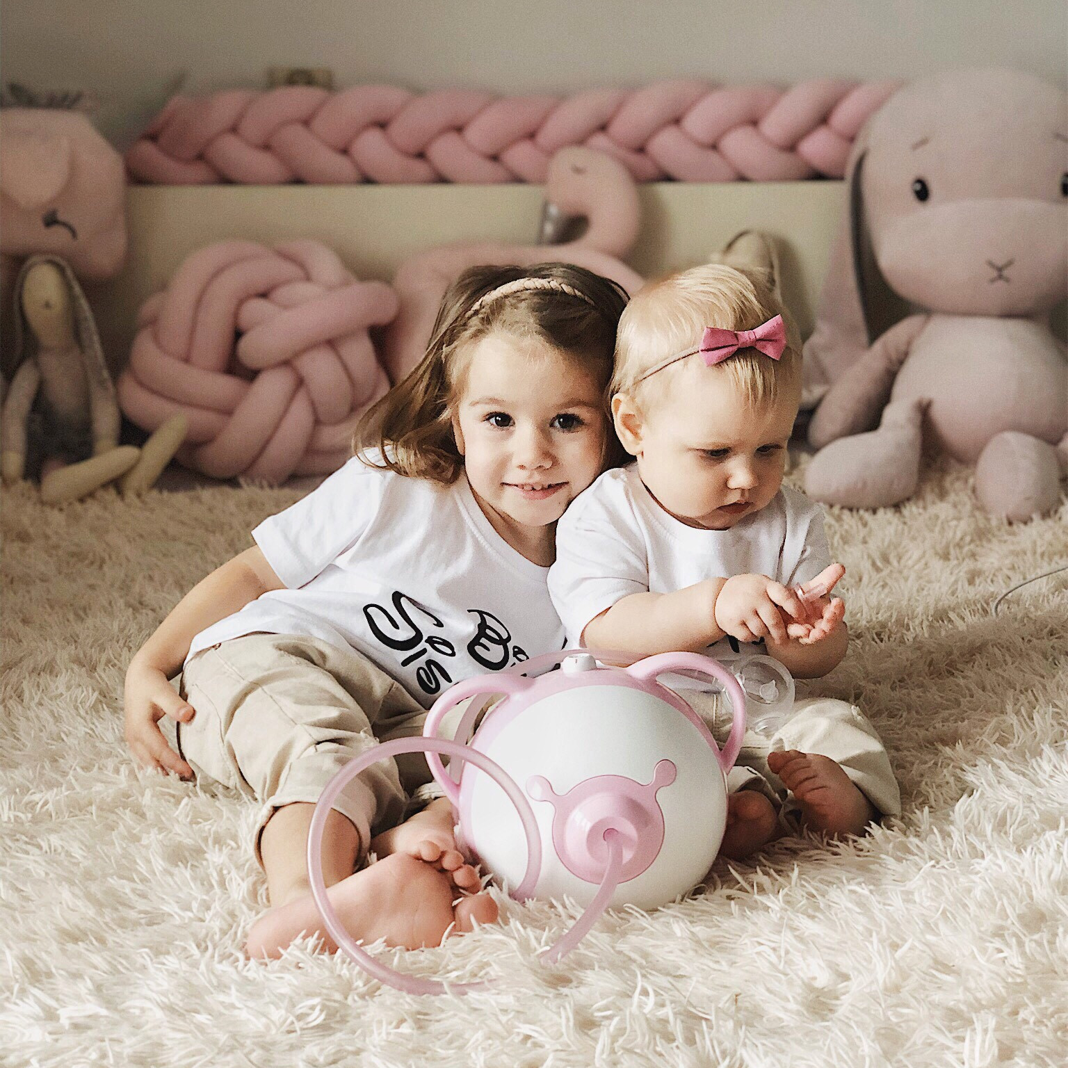Zwei hübsche kleine Mädchen sitzen nebeneinander auf einem Bett, vor ihnen der Nosiboo Pro elektrischer Baby Nasensauger in rosa Farbe