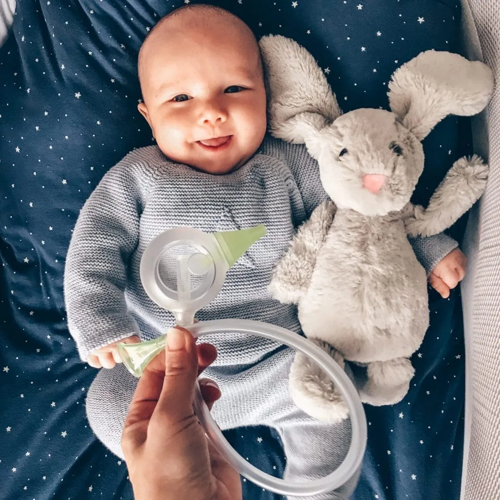 Im Vordergrund ist der manuelle Nasensauger Nosiboo Eco in der linken Hand einer Frau, im Hintergrund liegt ein lächelnd-süßes kleines Baby