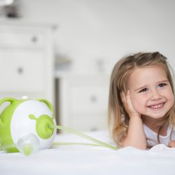 Öffnen Sie das Bild eines kleinen lächelnden Mädchens, das auf ihrem Bett neben dem Nosiboo Pro elektrischen Baby Nasensauger liegt